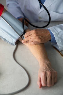 Erntehelfer mit Stethoskop, der an einem Tisch in einem medizinischen Raum sitzt und den Blutdruck mit einem Tonometer misst, während der Arbeit im Krankenhaus während des Ausbruchs des Coronavirus - ADSF14538