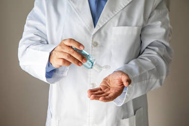 Crop male general practitioner in medical gown applying antibacterial gel for disinfecting of hands while standing on gray background in clinic during coronavirus pandemic - ADSF14539