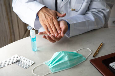 Male therapist in medical gown sitting at table in hospital and sanitizing hands with antiseptic while preparing for treatment of patients during coronavirus outbreak - ADSF14544