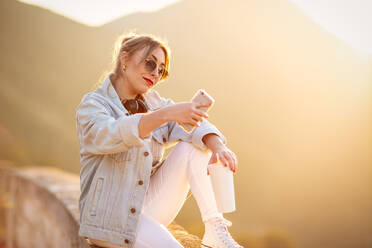 Nette Dame mit trendiger Sonnenbrille lächelnd bei der Aufnahme von Selfie auf Handy in warmen Licht der Sonne auf unscharfen Hintergrund - ADSF14587