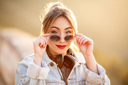 Fröhliche junge Frau mit Sonnenbrille in trendigem Freizeitoutfit lächelt und schaut in die Kamera an einem sonnigen Tag - ADSF14588