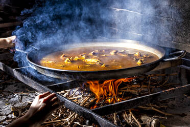 Crop unrecognizable hands holding big iron pan with boiling broth for cooking paella over open fire with wood - ADSF14596
