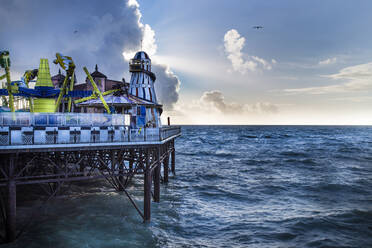 Colorful attractions of amusement park on pier near waving sea against cloudy sky in evening in Brighton, England - ADSF14599