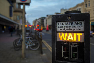 Modern instruction panel for traffic light with glowing wait sign on blurred background of evening street in Brighton, England - ADSF14600