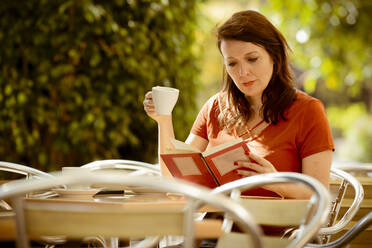 Erwachsene ruhige konzentrierte Dame lesen Buch und Tee trinken, während auf gemütlichen Sommer-Café Terrasse sitzen und genießen sonnigen Tag - ADSF14605