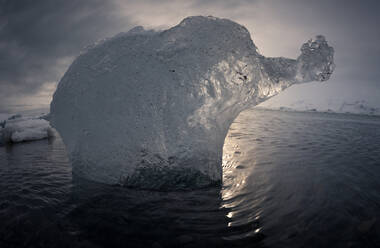 Durchsichtiger Eisberg, der im kalten Meerwasser gegen den bewölkten Abendhimmel in Island schwimmt - ADSF14621