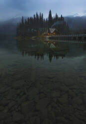 Friedliche Abendlandschaft mit ländlichen Haus mit beleuchteten Fenstern und Rauch aus der Pfeife am Ufer des ruhigen See mit klaren transparenten Wasser gegen dunklen Wald und nebligen Bergen in der kanadischen Landschaft gelegen - ADSF14626