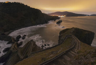 Wellen schlagen gegen eine mittelalterliche Steinbrücke, die zu einer kleinen Felseninsel mit der Einsiedelei San Juan de Gaztelugatxe auf der Spitze an der spanischen Küste führt, an einem sonnigen Tag mit blauem Himmel im Hintergrund - ADSF14631