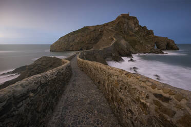 Wellen schlagen gegen eine mittelalterliche Steinbrücke, die zu einer kleinen Felseninsel mit der Einsiedelei San Juan de Gaztelugatxe auf der Spitze an der spanischen Küste führt, an einem sonnigen Tag mit blauem Himmel im Hintergrund - ADSF14632