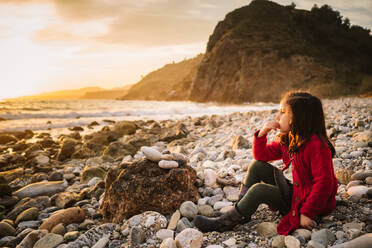 Seitenansicht eines nachdenklichen weiblichen Kindes, das am steinigen Strand sitzt und den Sonnenuntergang vor dem Hintergrund eines ruhigen Meeres bewundert - ADSF14656