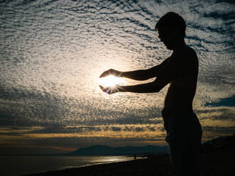 Silhouette eines Mannes, der die Sonne mit einer Geste hält, vor dem Hintergrund eines malerischen bewölkten Sonnenuntergangs - ADSF14658