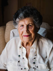 Portrait of happy senior curly gray haired woman in white shirt and with beads on neck looking at camera at home - ADSF14660