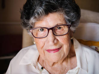 Lächelnde, ältere, lockige, grauhaarige Frau mit Brille und rosa Lippen, die in einer Wohnung in die Kamera schaut - ADSF14663