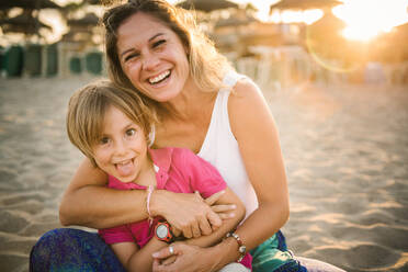 Beautiful laughing woman embracing cute boy showing tongue while sitting together on beach in bright sunshine - ADSF14668