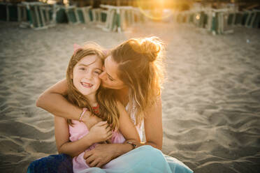 Adult woman kissing beautiful girl embracing with love on sandy beach in sunset light - ADSF14670