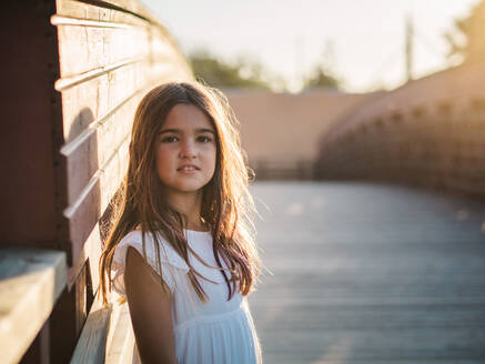 Porträt eines niedlichen kleinen Mädchens im weißen Kleid, das im Sonnenlicht an der Wand steht und in die Kamera schaut - ADSF14676