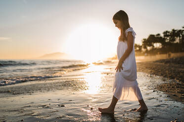 Seitenansicht eines kleinen Mädchens in einem weißen Kleid, das am Meeresufer spazieren geht, vor dem Hintergrund der Sonne - ADSF14677