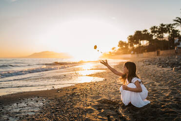 Side view of little girl in white dress sitting on beach and playing with stones on background of sundown - ADSF14678