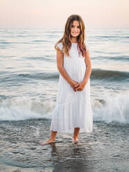 Portrait of charming little girl in white dress standing in water on sandy beach and looking at camera - ADSF14680