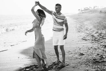 Full body adult man smiling and spinning woman in dance while having fun on sandy beach near sea - ADSF14682