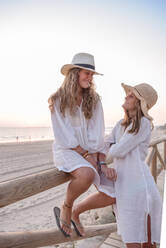 Charming relaxed women in white dresses and hat smiling while speaking comfortable placing on wooden fence on sandy beach in sunny day - ADSF14685