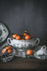 Sophisticated still life with ripe orange tangerines with green leaves in deep white bowl on table on gray background - ADSF14696
