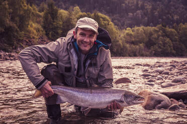 Fly fisherman smiling while holding caught salmon fish in river - DHEF00357