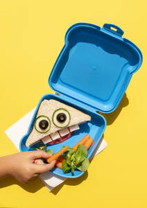 Hand of baby girl picking up baby carrot from lunch box with funny looking sandwich with anthropomorphic face - GEMF04134