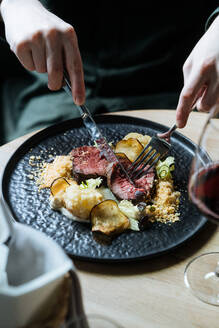 Bildausschnitt von oben von Person mit schwarzem Teller mit Roastbeefscheiben auf Sahnesauce mit Blumenkohl und gebratenen Pommes frites beim Abendessen mit einem Glas Rotwein - ADSF14736