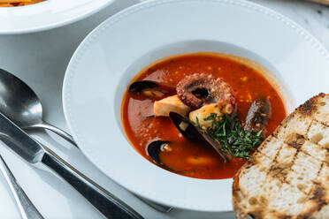 Draufsicht auf eine rote Suppe mit Tintenfisch und Muscheln, serviert mit Grünzeug und geröstetem Brot im Restaurant - ADSF14748