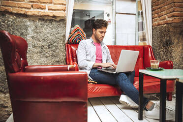 Mittlerer erwachsener Mann, der einen Laptop benutzt, während er auf dem Sofa in einem Café sitzt - EHF00792