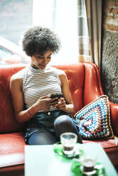 Woman with curly hair using smart phone while relaxing on sofa against window in cafe - EHF00793