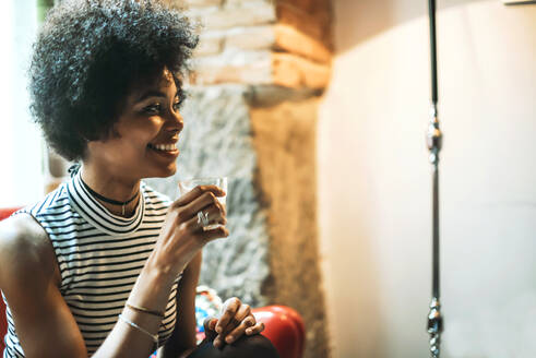 Nahaufnahme einer lächelnden Frau mit lockigem Haar, die einen Kaffee in der Hand hält und an der Wand eines Cafés sitzt - EHF00795