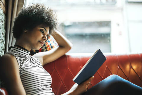 Mittlere erwachsene Frau mit lockigem Haar liest ein Buch und entspannt sich auf dem Sofa in einem Café, lizenzfreies Stockfoto