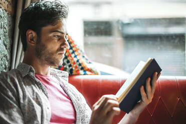 Nahaufnahme eines ernsten, erwachsenen Mannes, der ein Buch liest, während er in einem Café am Fenster sitzt - EHF00800