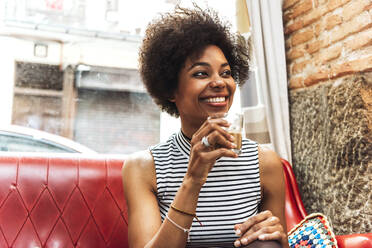 Nahaufnahme einer lächelnden Frau mit lockigem Haar, die Kaffee trinkt, während sie auf einem Sofa in einem Café sitzt - EHF00804