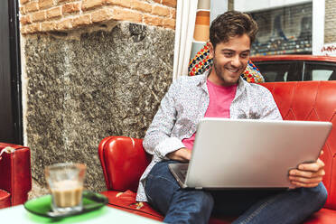 Lächelnder mittelgroßer erwachsener Mann, der einen Laptop benutzt, während er auf dem Sofa im Cafe sitzt - EHF00807