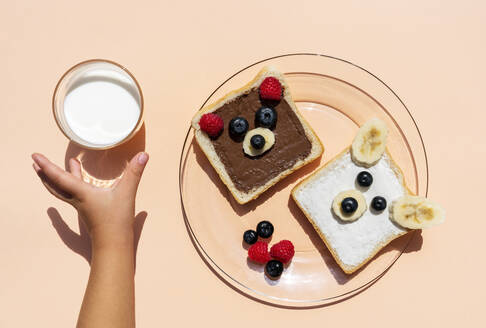 Studio shot of toasts with bear faces made of fruits and hand of baby girl reaching for glass of milk - GEMF04139