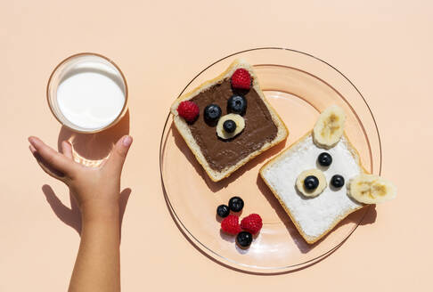 Studioaufnahme von Toasts mit Bärengesichtern aus Früchten und der Hand eines kleinen Mädchens, das nach einem Glas Milch greift - GEMF04139