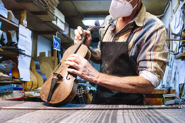 Craftsman with face mask examining string of violin on workbench at workshop - JCMF01214