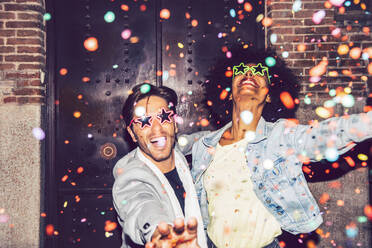 Confetti falling on cheerful couple wearing star shaped sunglasses enjoying outdoors at night - EHF00865