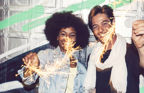 Fröhliches Paar spielt mit Wunderkerzen, während es nachts an der Wand steht, lizenzfreies Stockfoto