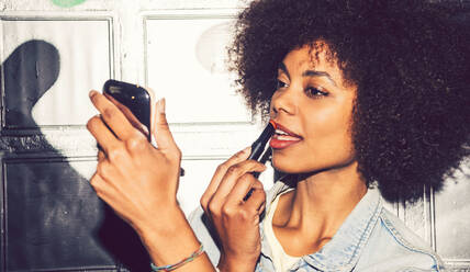 Close-up of woman with curly hair applying lipstick while looking in mirror at night - EHF00887