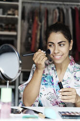Smiling young woman applying blusher on face while looking at mirror - JMHMF00095