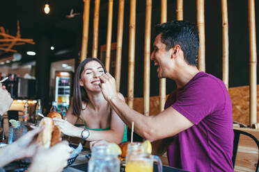 Cheerful young man giving snack to laughing girlfriend while having lunch with friends in cafeteria - ADSF14769