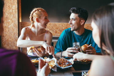 Happy young man and woman talking and laughing while gathering with friends in cafeteria and enjoying fast food snacks and drinks - ADSF14770