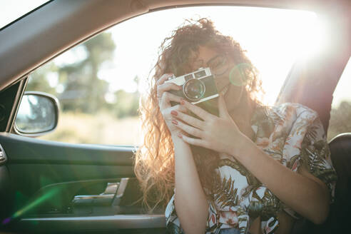 Zufriedene Frau in Freizeitkleidung und mit Brille im Auto sitzend, während sie ein Foto mit einer Digitalkamera im Sonnenlicht im Gegenlicht aufnimmt - ADSF14772