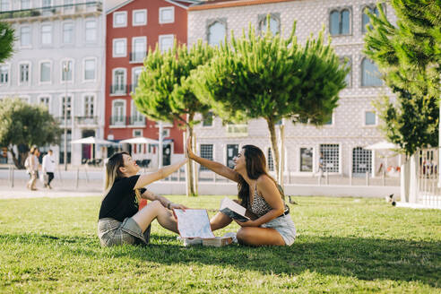 Weibliche Touristen geben sich High-Five, während sie auf einer Wiese vor einem Gebäude im Park sitzen - DCRF00779