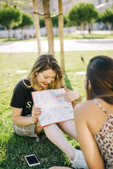 Smiling tourist showing map to female friend while sitting on grassy land in park - DCRF00781