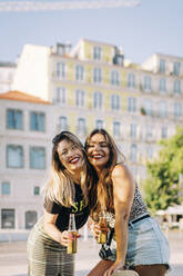 Fröhliche Freundinnen halten Bierflaschen, während sie an einem Gebäude in der Stadt stehen - DCRF00788
